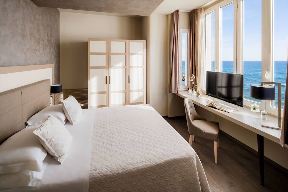 A hotel room with a bed, desk, TV, and a view of the sea.