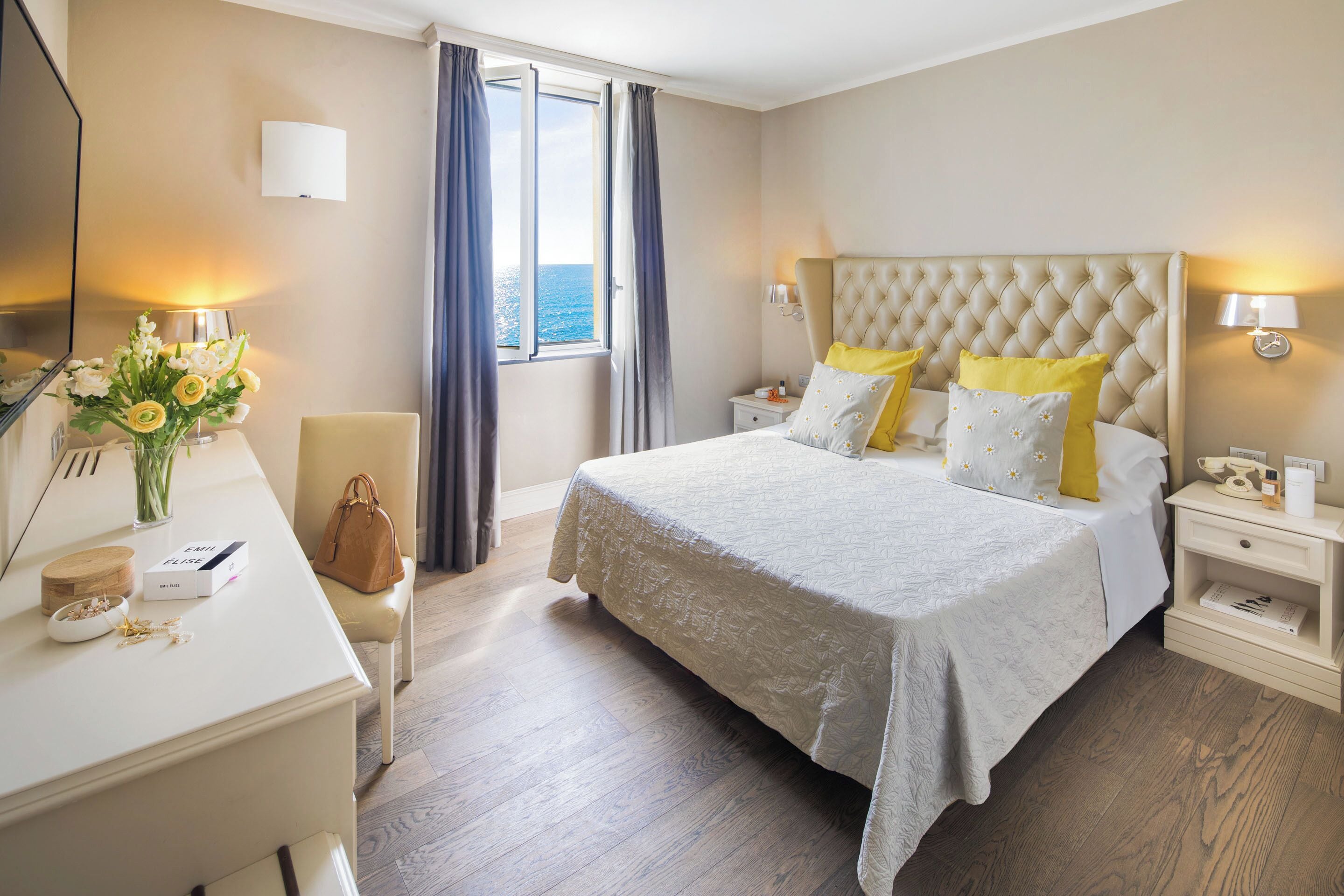 Habitación de hotel con una cama grande, mesitas de noche, un escritorio con una silla, un florero con flores y una ventana con cortinas.