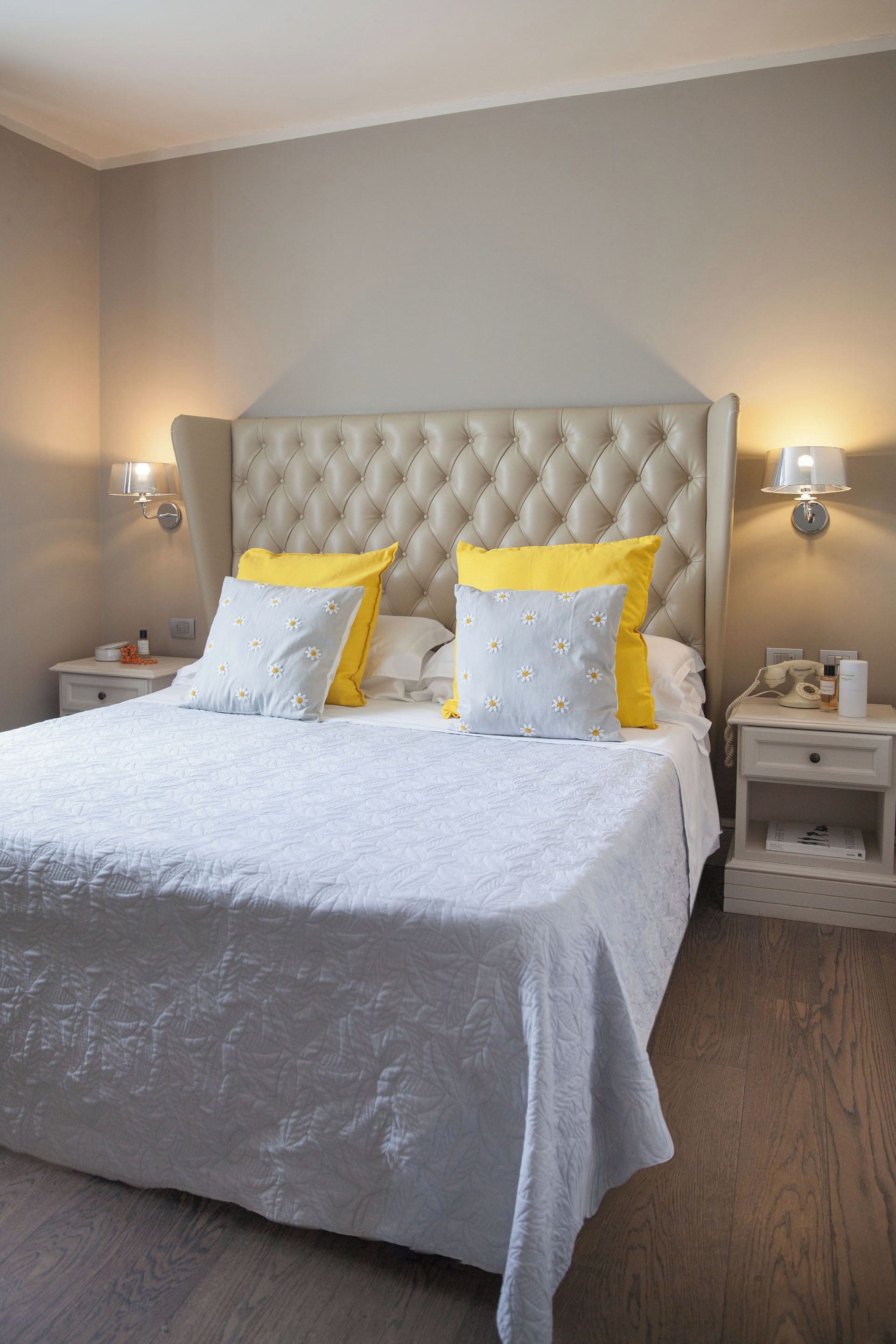 Une chambre à coucher avec une tête de lit capitonnée, un lit avec une literie blanche et des oreillers jaunes, des tables de chevet avec des lampes et un plancher en bois.