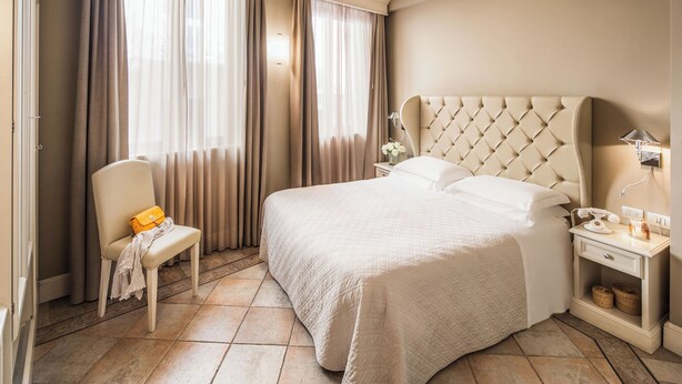 A neatly arranged hotel room with a large bed, a chair, a nightstand, and curtains. - Hotel Savoia (Alassio)
