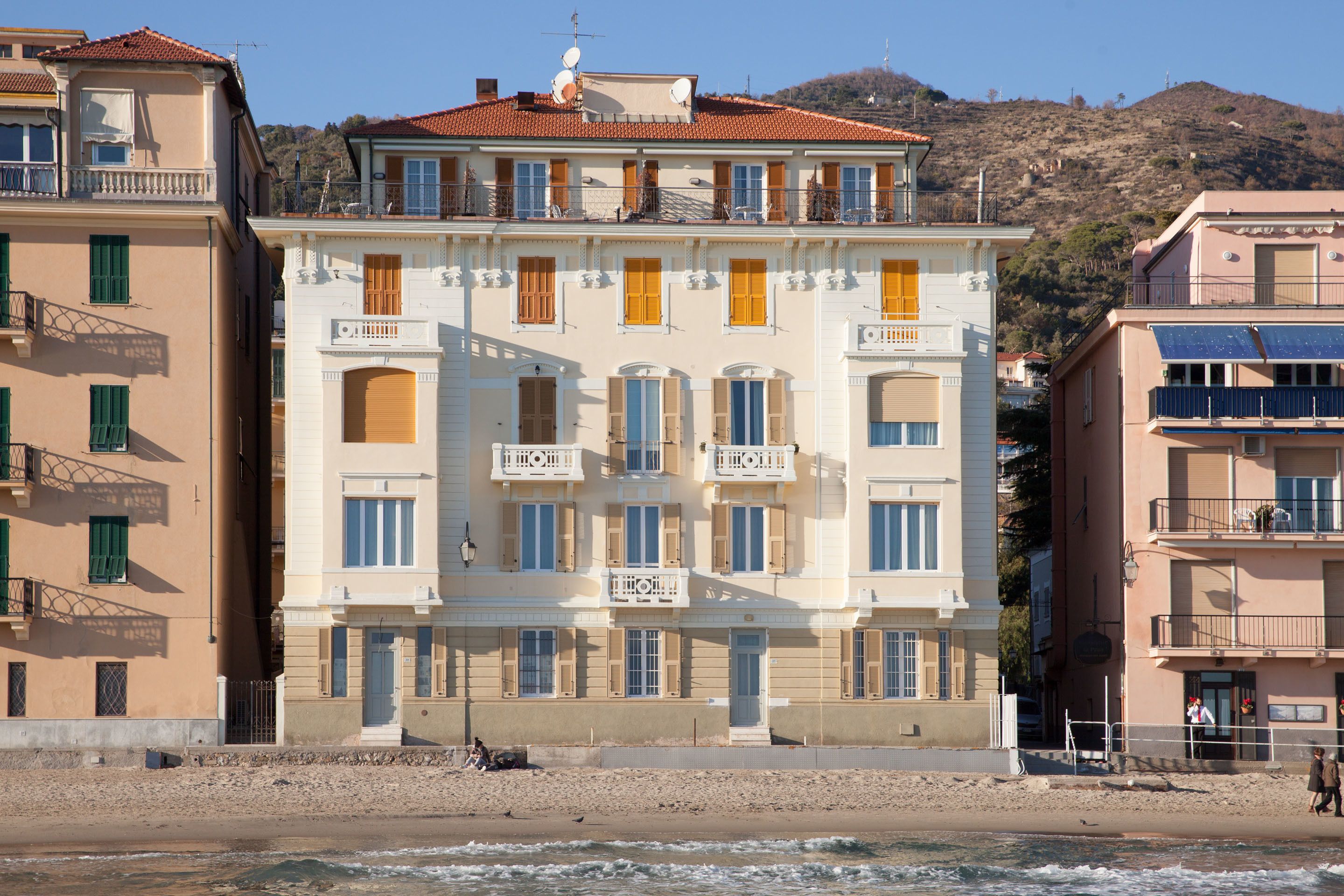 A multi-story building with balconies, a beach in the foreground, and a hilly landscape in the background.