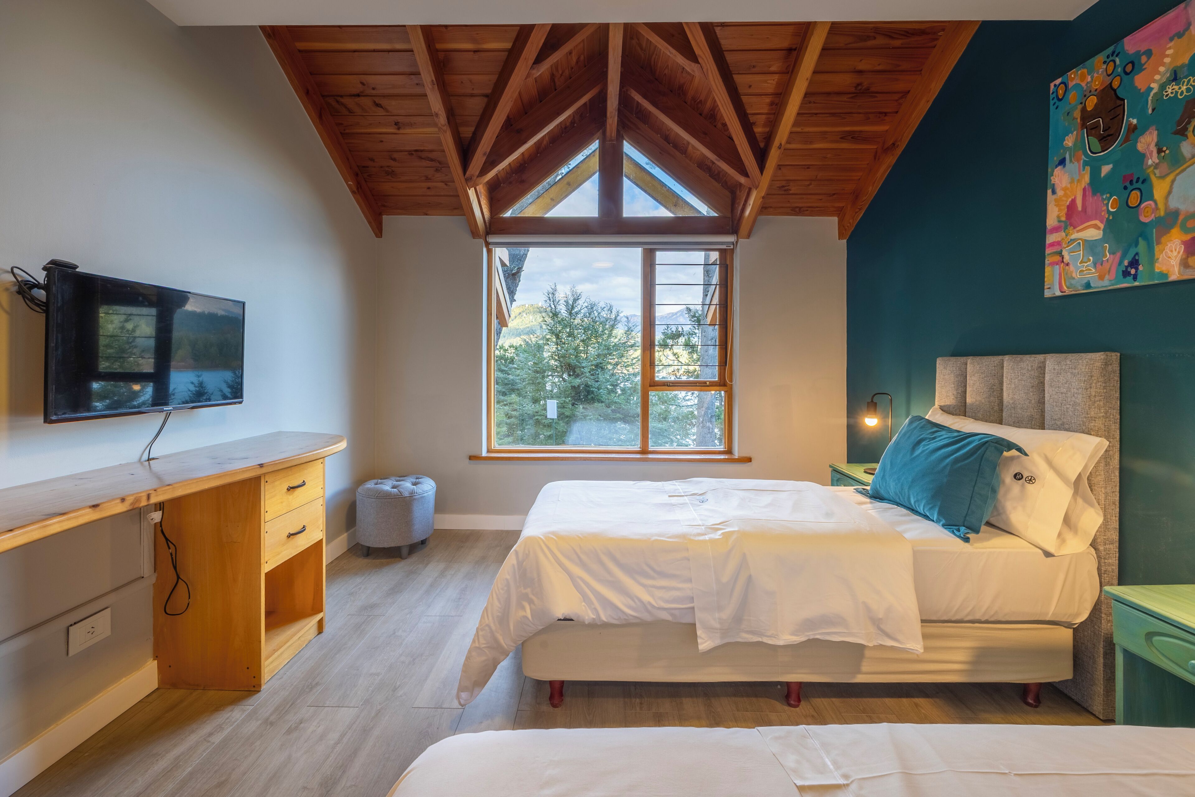 Un dormitorio con cama, escritorio, televisor y ventana con vista a árboles.