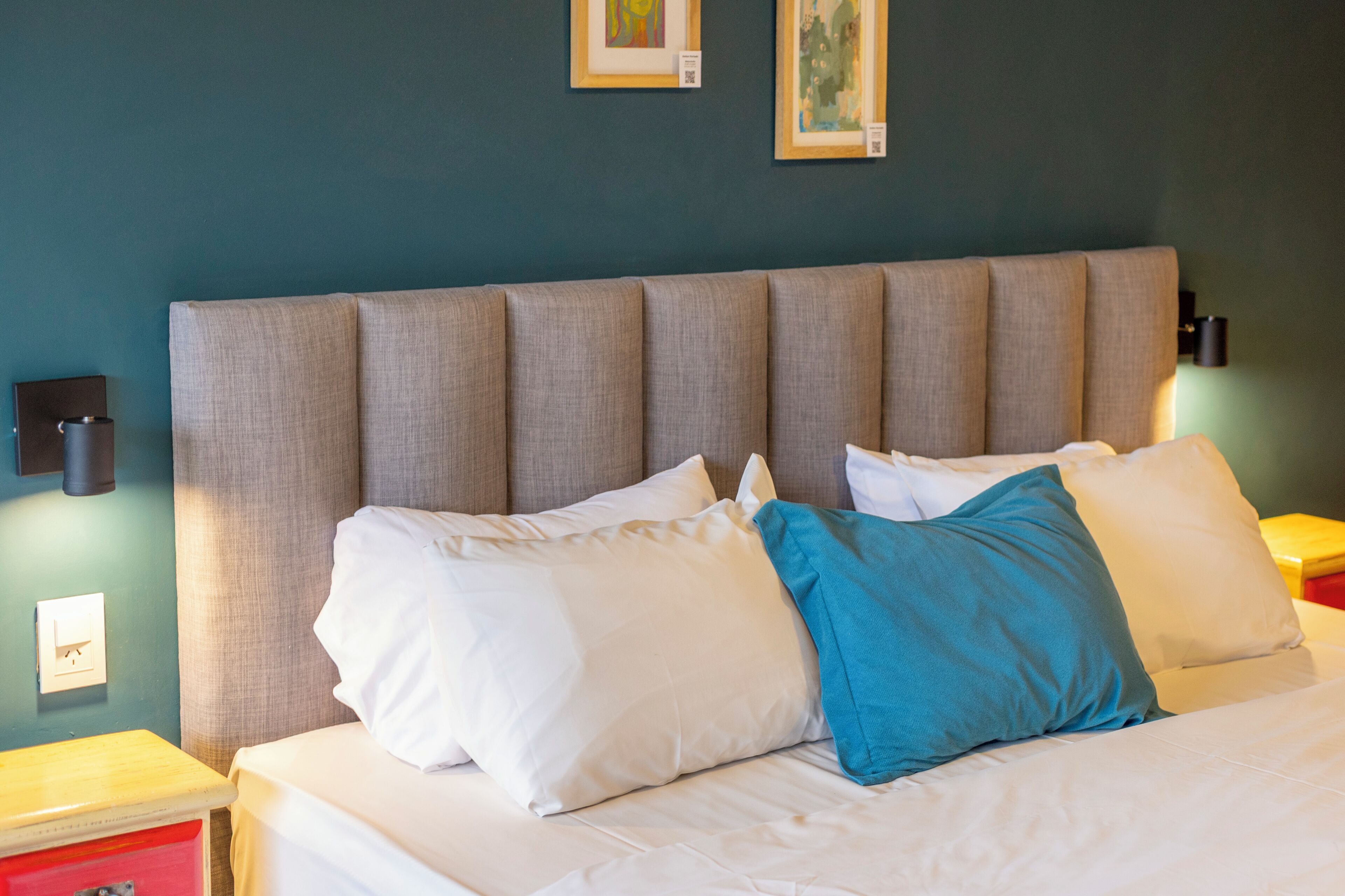 A bed with white and blue pillows, a grey headboard, and a red nightstand with a lamp.