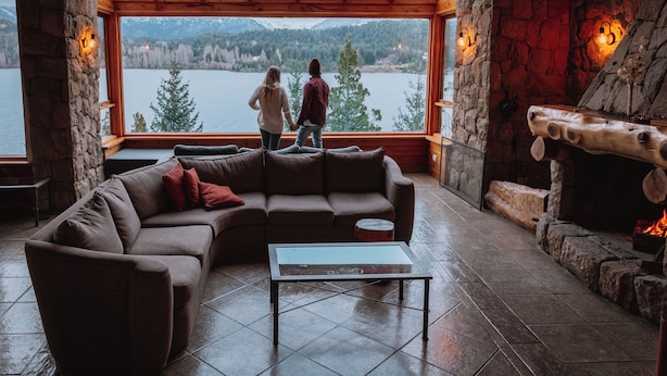 Un interior acogedor con una chimenea de piedra, un amplio ventanal con vista a un lago y un sofá modular.
