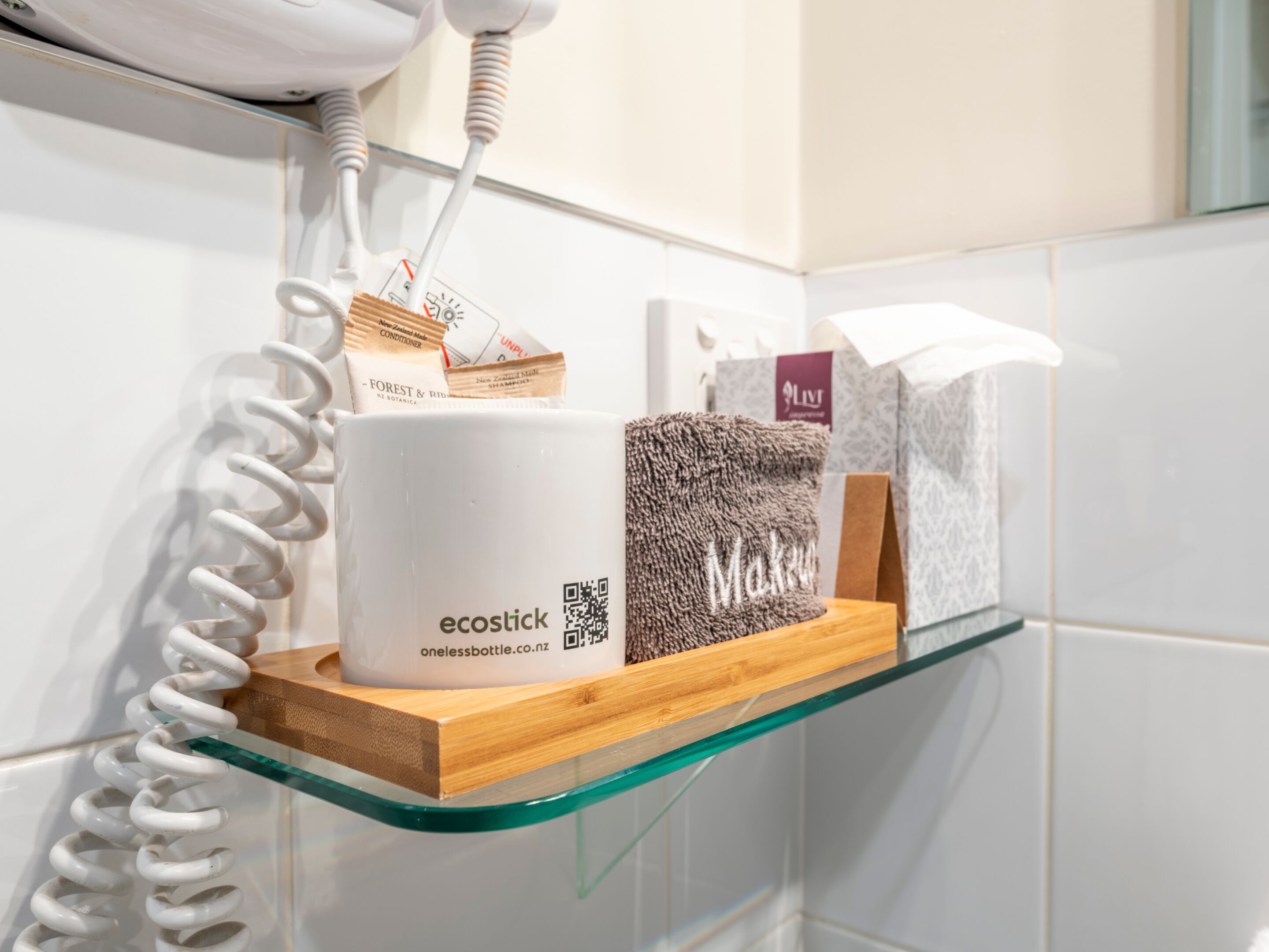 Un baño con un estante de vidrio que sostiene una taza blanca, una toalla marrón y una caja con la etiqueta 'Makeup'.