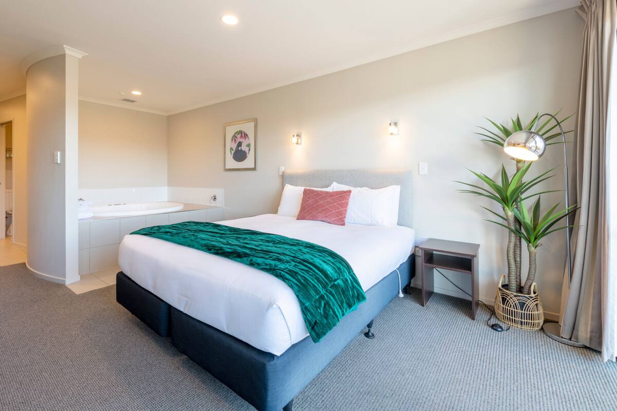 A modern hotel room with a large bed, a built-in bathtub, a small bedside table, and a potted plant.