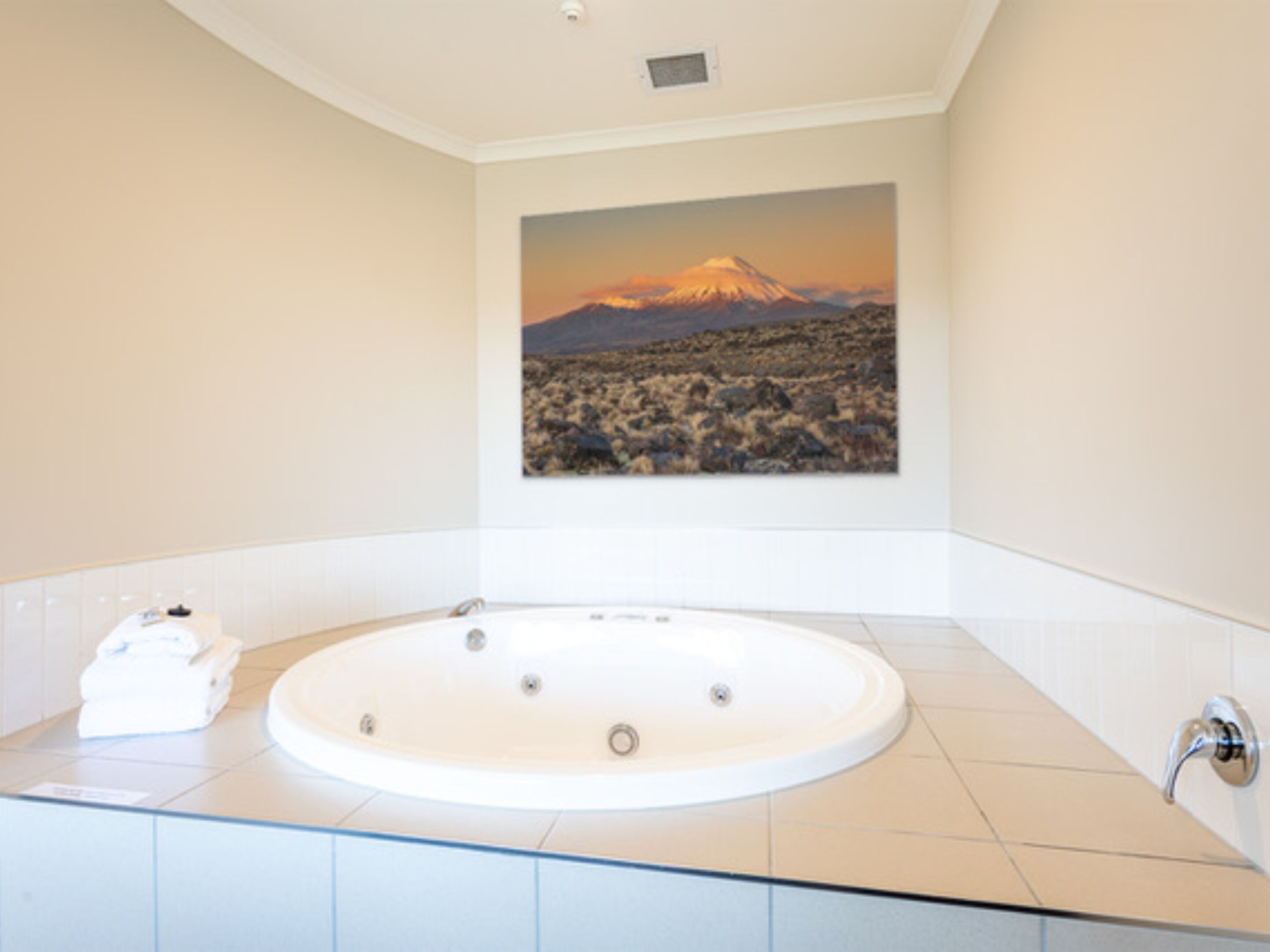 A bathroom with a large bathtub, white tiles, and a painting of a mountain landscape.