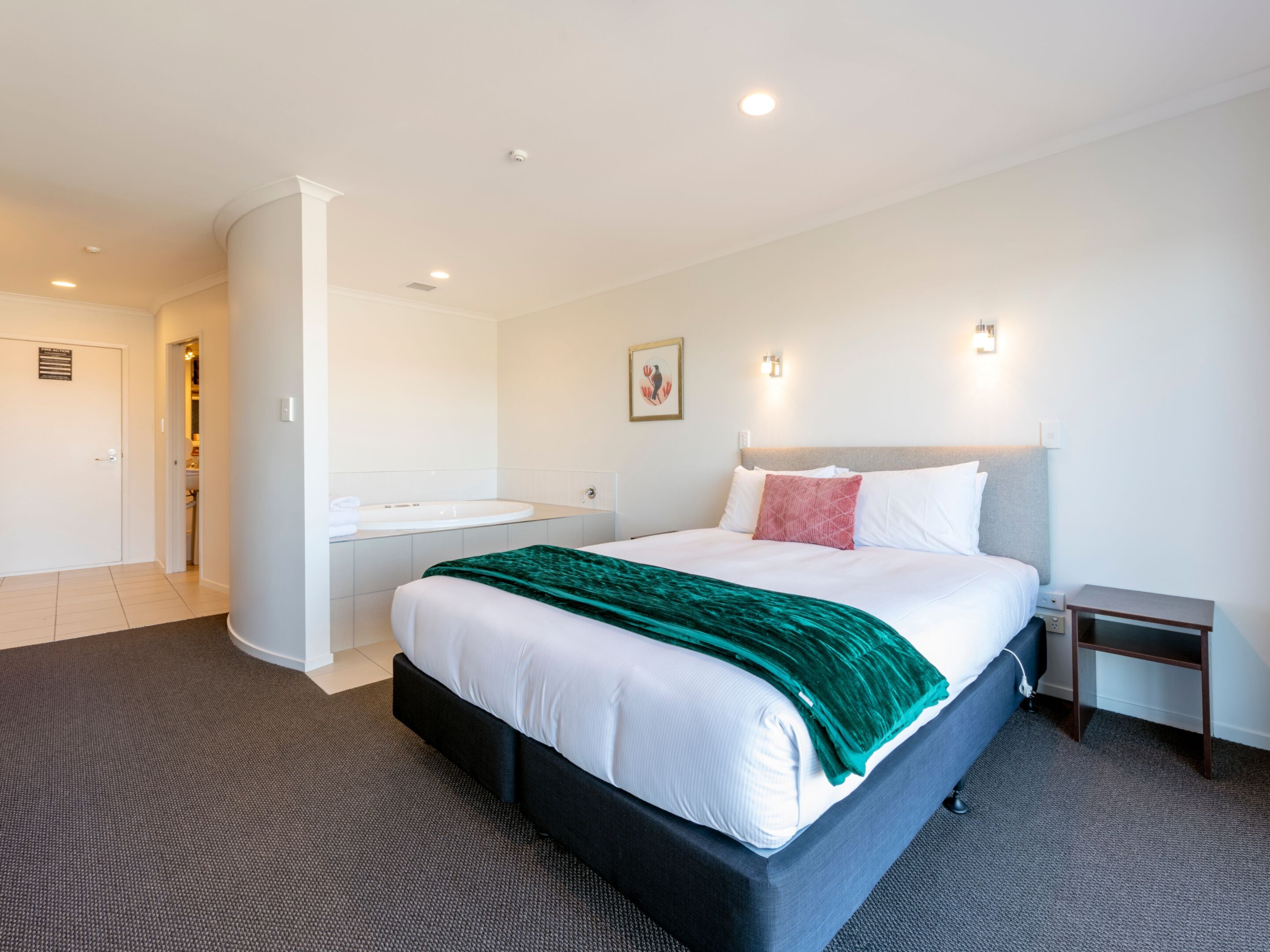 A hotel room with a large bed, a bedside table, a nightstand, a bathroom with a sink and mirror, and a door leading to another room.