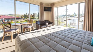A hotel room with a large bed, a desk, two chairs, a TV on the wall, and a balcony with a view of the city. - Lake Taupo Motor Inn (Taupo)