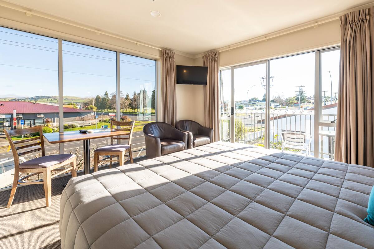 A hotel room with a large bed, a desk, two chairs, a TV on the wall, and a balcony with a view of the city.