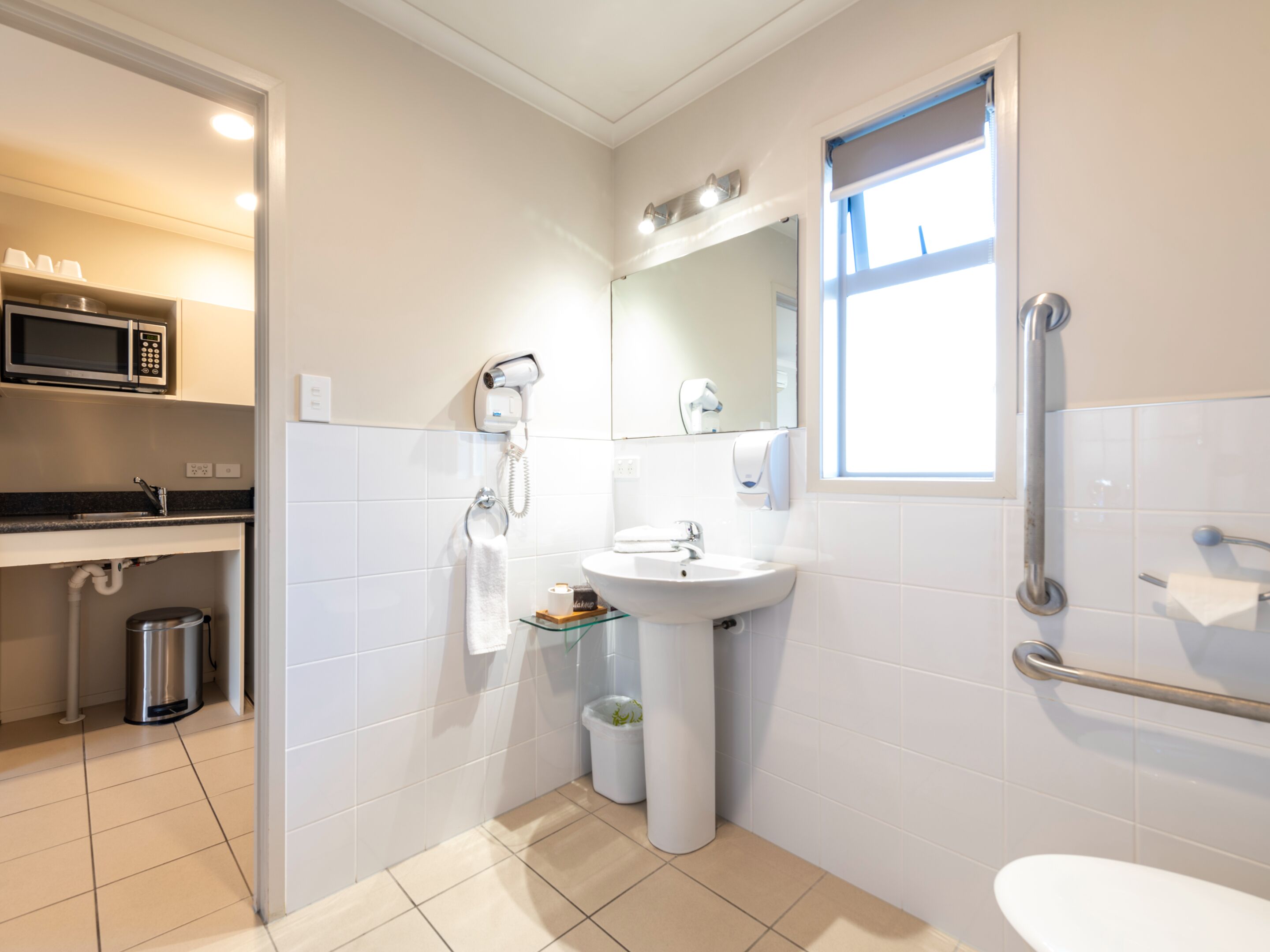 A modern bathroom with a sink, mirror, and toilet. There is a window providing natural light, a hairdryer, and a trash bin.