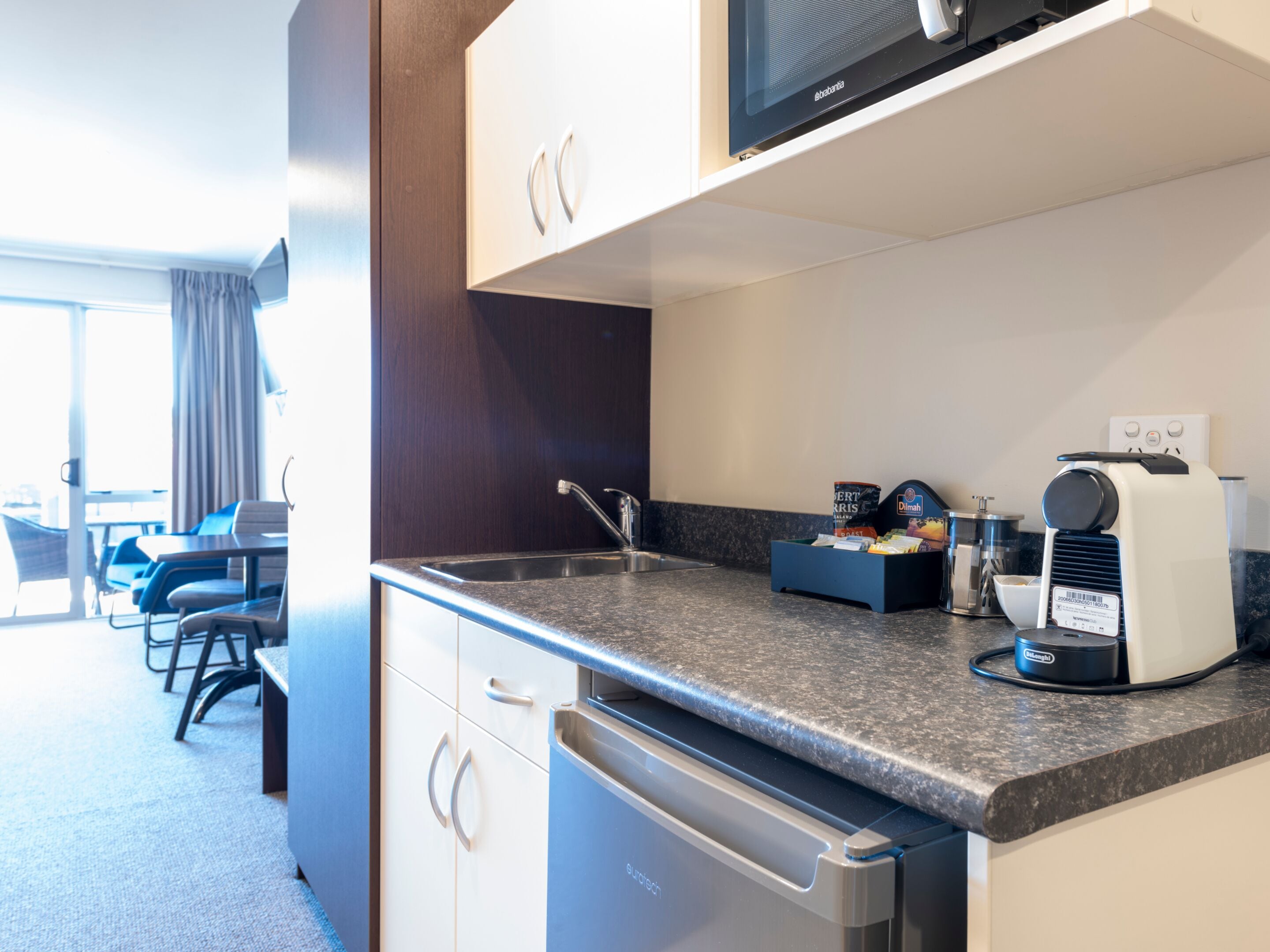 A modern kitchen with a built-in dishwasher, a coffee machine, and a microwave. There is a small seating area with a table and chairs visible in the background.