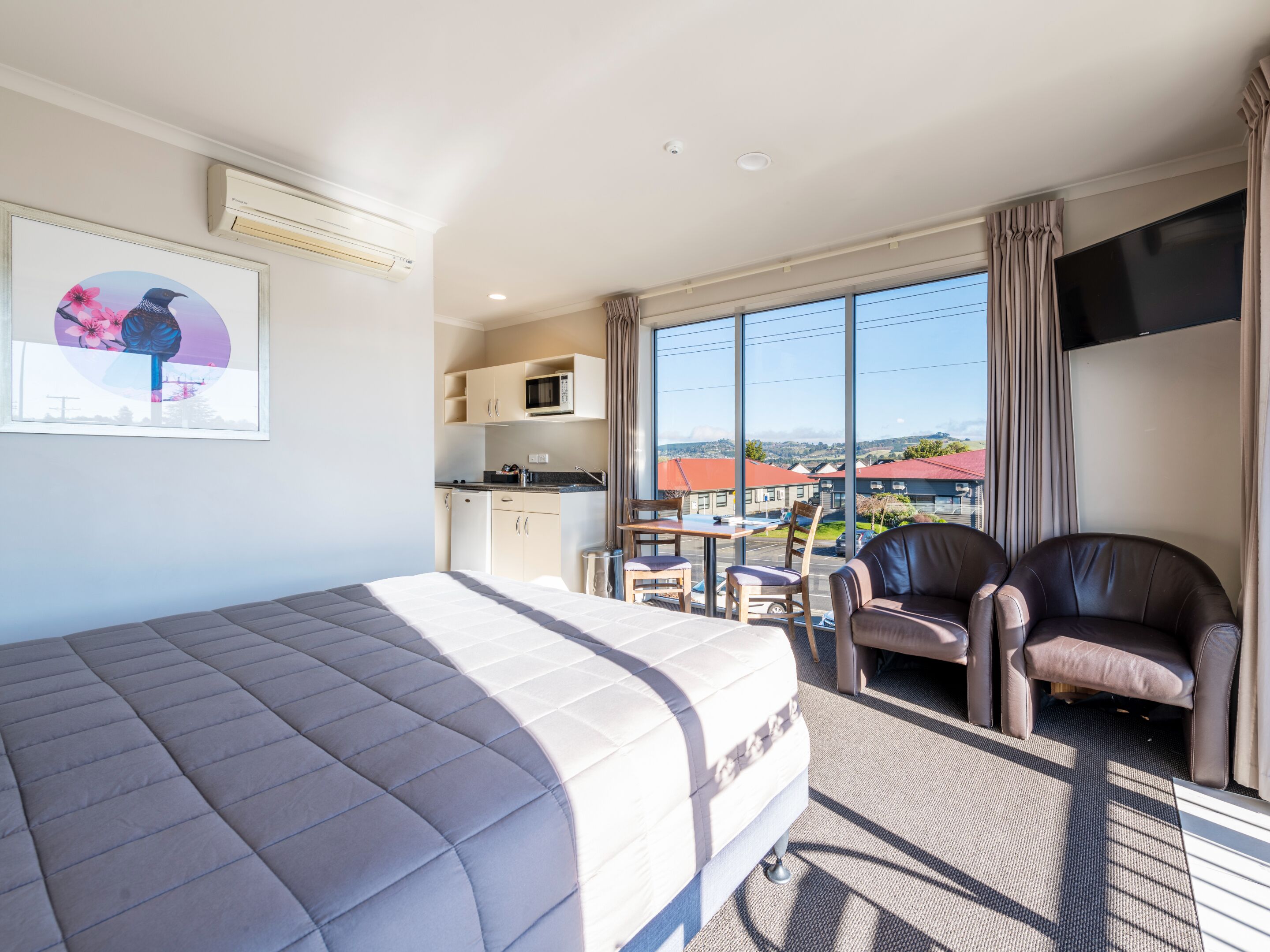 Quarto de hotel com uma cama, uma escrivaninha, duas cadeiras, uma TV e uma janela grande com vista para edifícios.