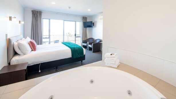A modern hotel room with a large bed, a sitting area with a chair, a flat-screen TV mounted on the wall, and a large bathtub. - Lake Taupo Motor Inn (Taupo)