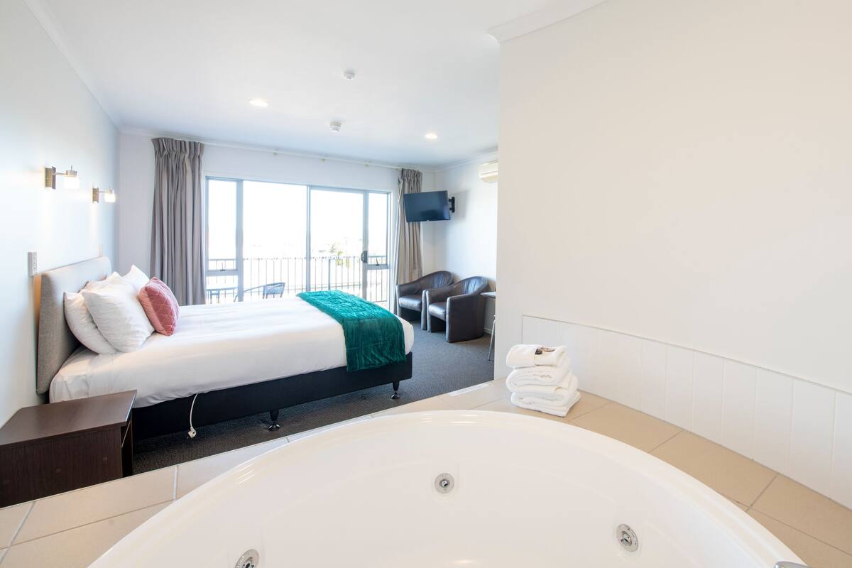 A modern hotel room with a large bed, a sitting area with a chair, a flat-screen TV mounted on the wall, and a large bathtub.