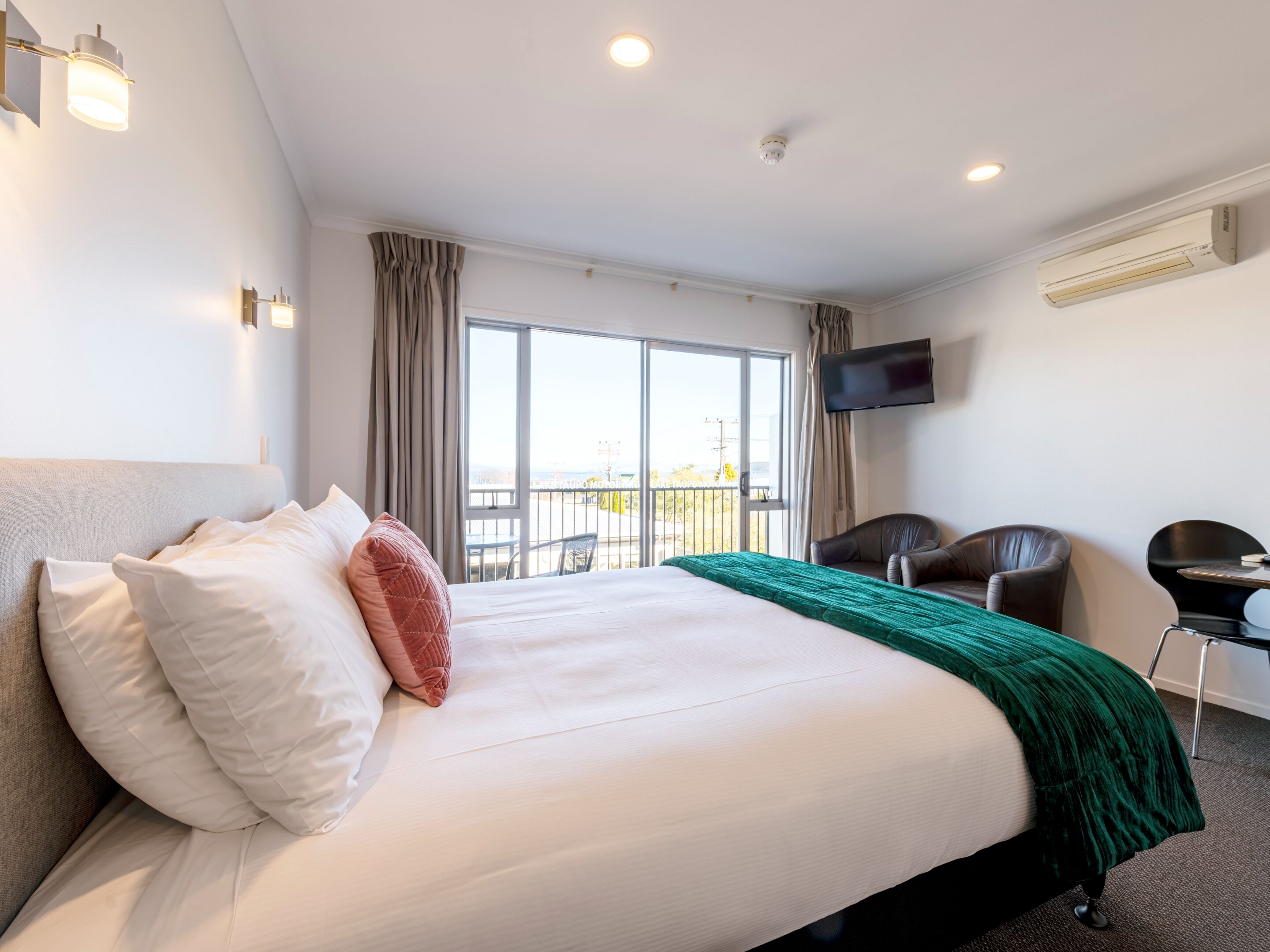 A hotel room with a large bed, a TV mounted on the wall, a balcony with a view, and a desk with a chair.
