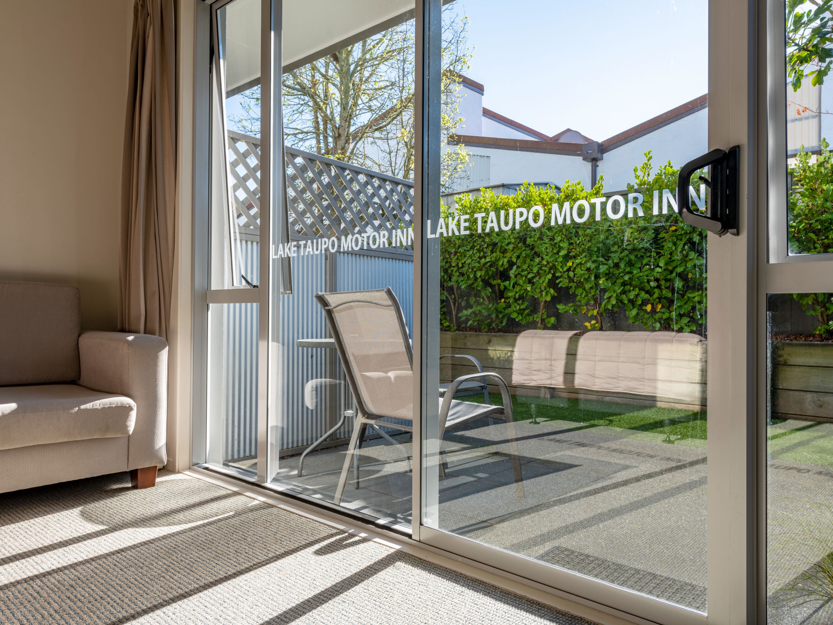 A modern living room with a sofa, a chair, and a glass door leading to a patio with a pool and greenery.