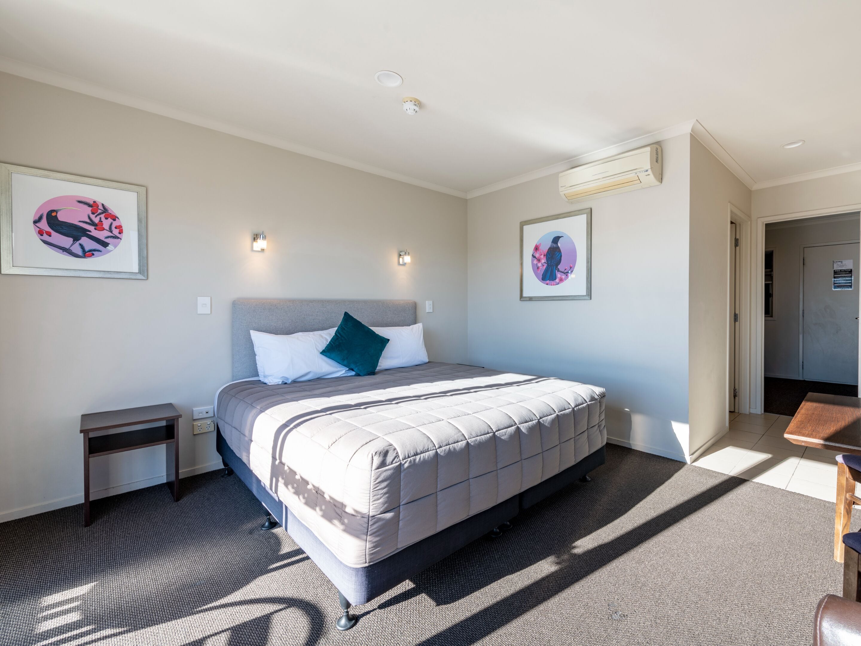 Ein Hotelzimmer mit einem großen Bett, zwei Nachttischen, einem kleinen Schreibtisch und einem Sessel. An den Wänden befinden sich zwei Wandleuchten und zwei gerahmte Bilder.