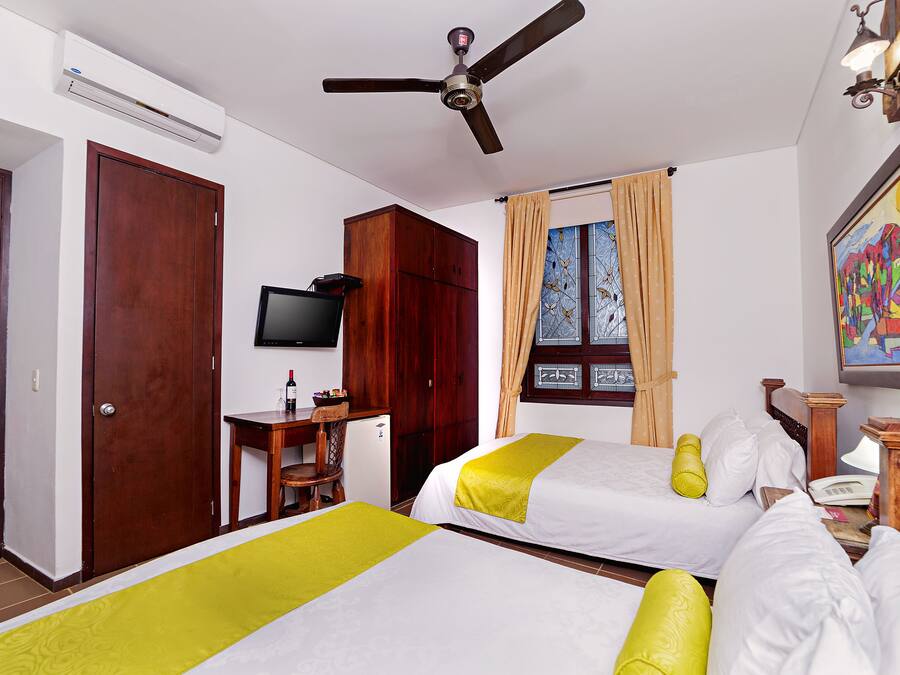A hotel room with two beds, a ceiling fan, an air conditioner, a desk with a chair, a television, and a window with curtains.