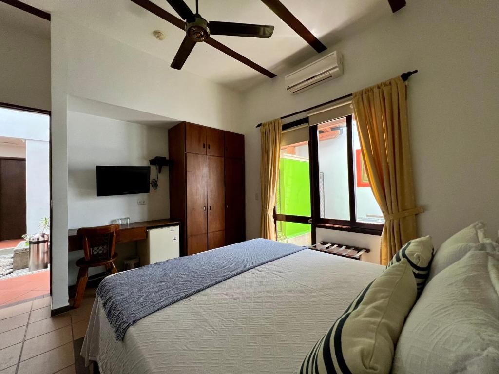 Habitación de hotel con cama, escritorio, silla, televisor, ventilador de techo y ventana con cortinas.