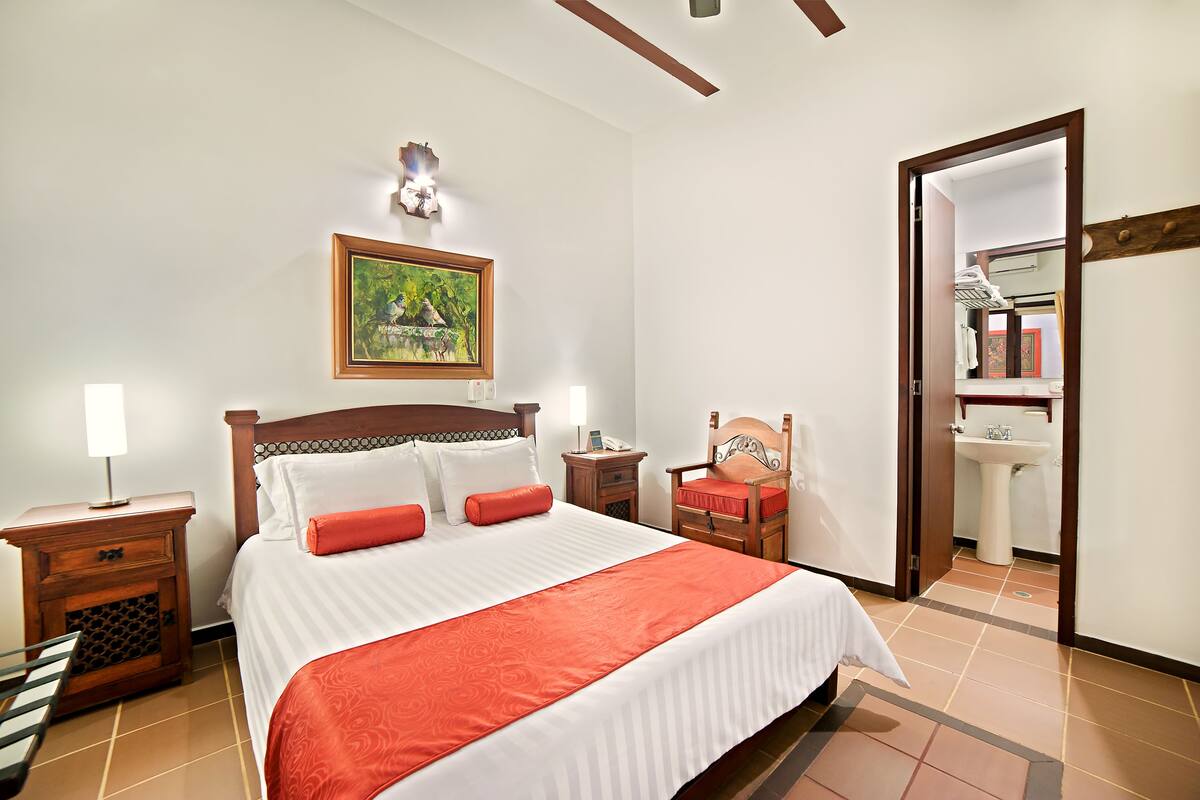 Ein ordentlich eingerichtetes Hotelzimmer mit Bett, Nachttischen, einem Sessel, einem Bild und einem durch eine offene Tür einsehbaren Badezimmer.