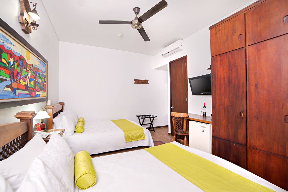 Ein Hotelzimmer mit zwei Betten, einem Deckenventilator, einem Schreibtisch mit Stuhl, einem Fernseher und einem Kleiderschrank.