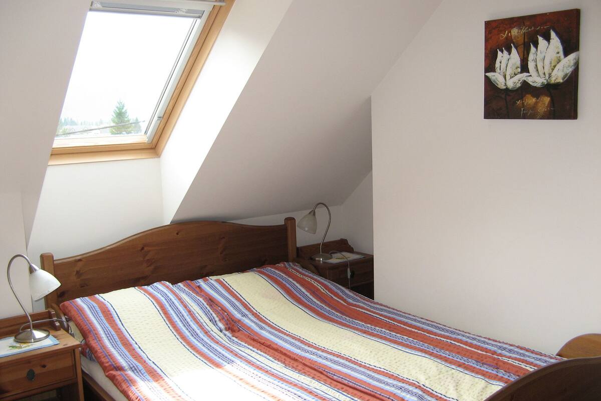 Une chambre avec un plafond en pente, un lit en bois, une table de chevet avec une lampe, une commode et une fenêtre.