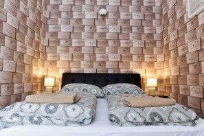 A room with a brick-patterned wall, a black upholstered headboard, two bedside tables with lamps, and patterned bedding. - Corvin Point Rooms and Apartments (Budapest)