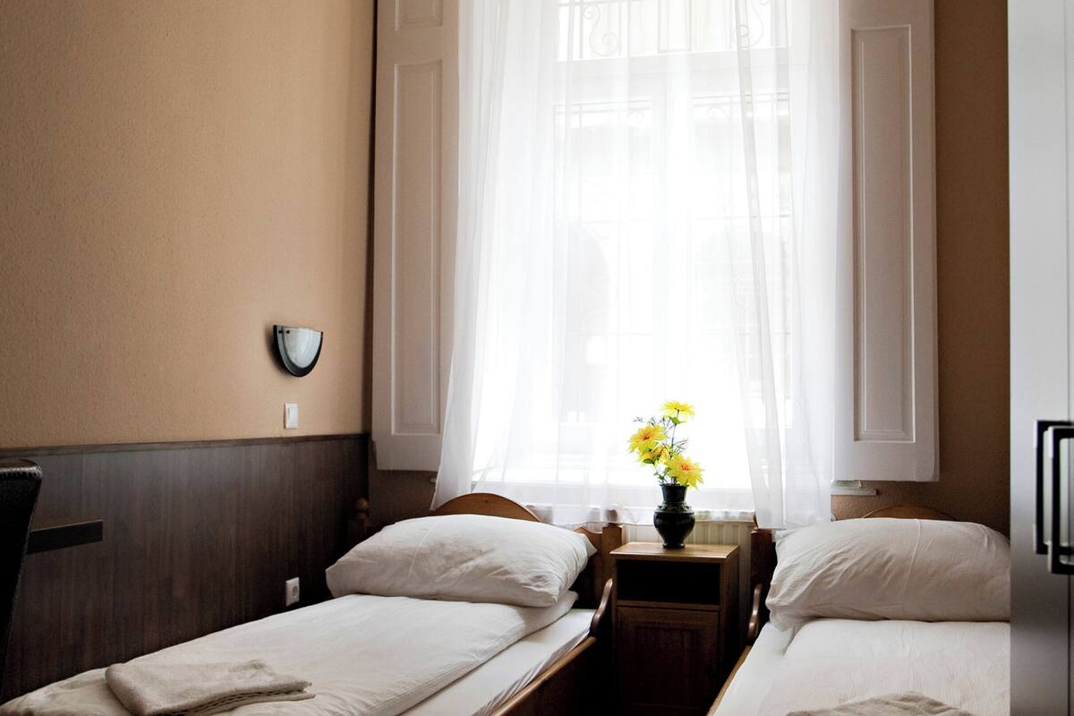 A hotel room with two single beds, a bedside table with a vase of flowers, a wall-mounted lamp, and a window with sheer curtains.