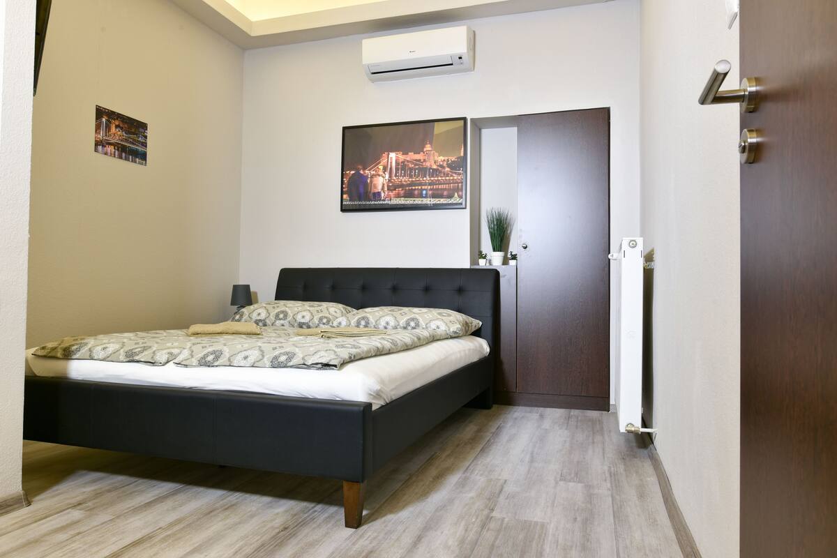 A bedroom with a bed, a door, a wall-mounted air conditioner, and a framed picture.