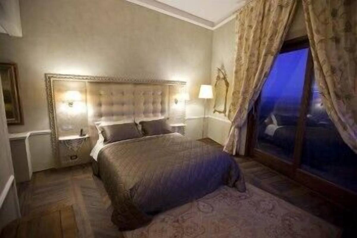 A bedroom with a large bed, a headboard, two bedside lamps, and a window with curtains.