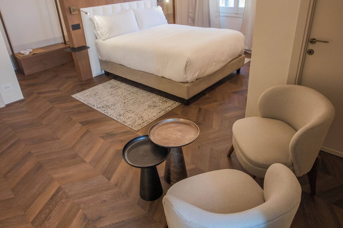 A modern hotel room with a large bed, a sitting area with two chairs, a small table, and a herringbone wood floor.