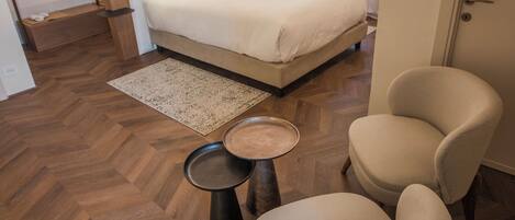 Una habitación de hotel moderna con una cama grande, una zona de estar con dos sillas, una mesita y un suelo de madera con estampado de espiga.