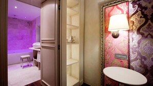 Een badkamer met een wastafel, een wasbak en een spiegel. Er is een plankje met een beker en een zakdoos. De muren zijn versierd met behang met een patroon.