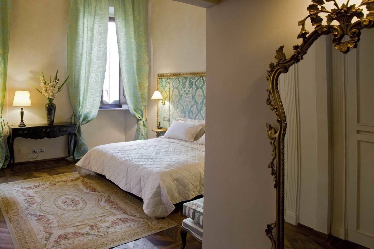 A bedroom with a bed, bedside tables, a lamp, a chair, a table, and a window with curtains.