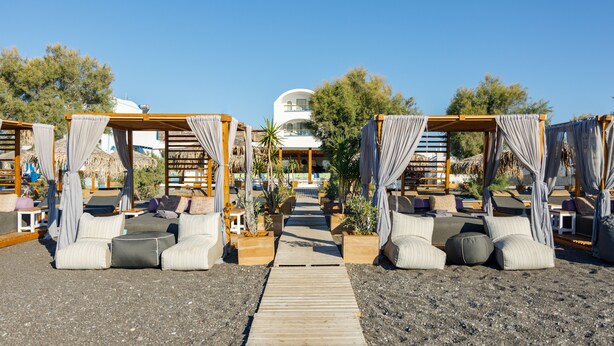 A beachside resort with thatched-roof cabanas, cushioned seating, and a wooden pathway leading to a building. - Glykeria Mini Suites (Santorini)