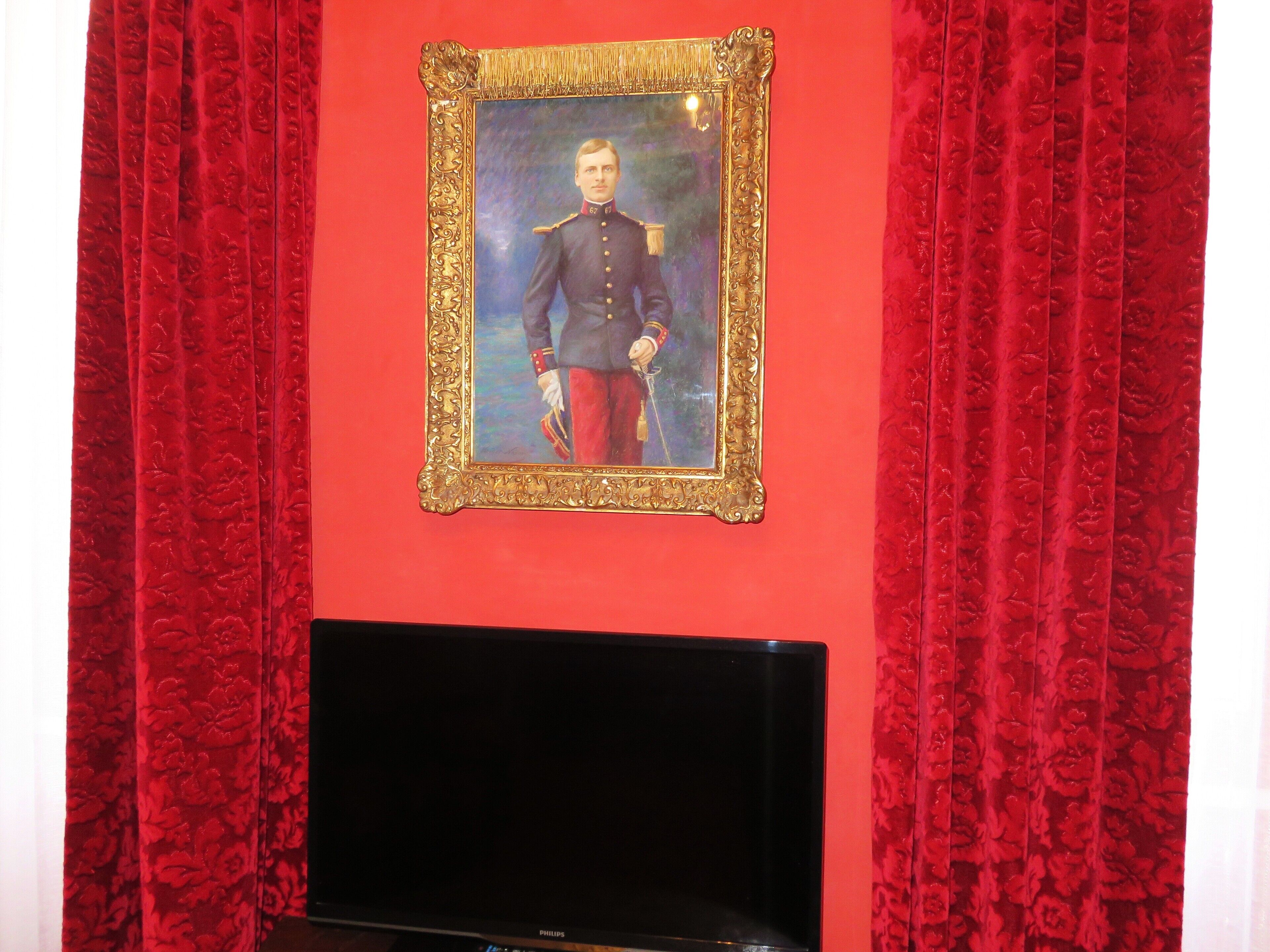 A room with a portrait framed in gold, red curtains, and a black television on a stand.