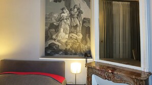 A hotel room with a large wall mural, a bed with a red accent, a nightstand with a lamp, and a mirror above the bed. - Hôtel Windsor Home (Paris)