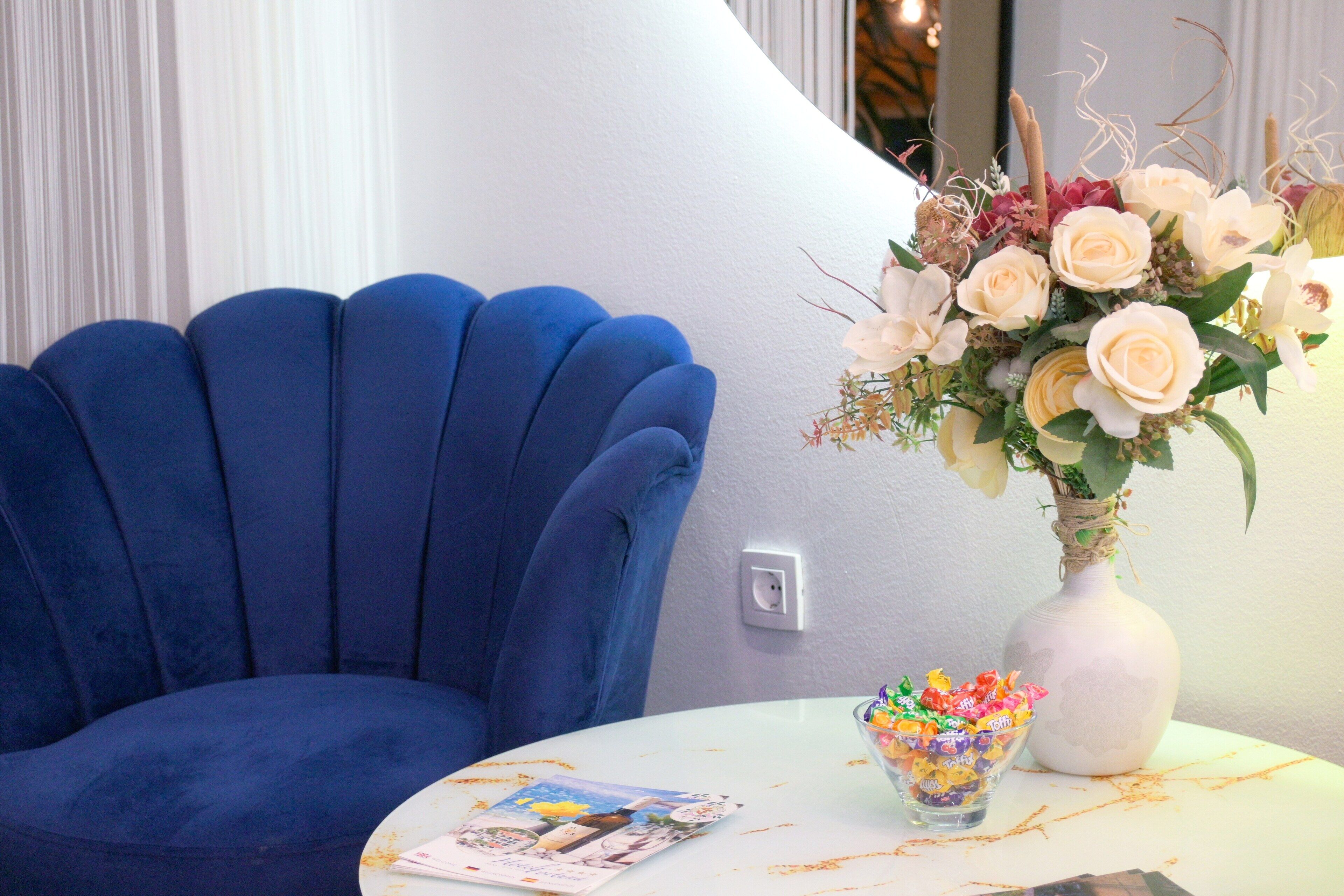 Una silla tapizada azul, un jarrón blanco con flores y un cuenco con caramelos de colores sobre una mesa redonda.