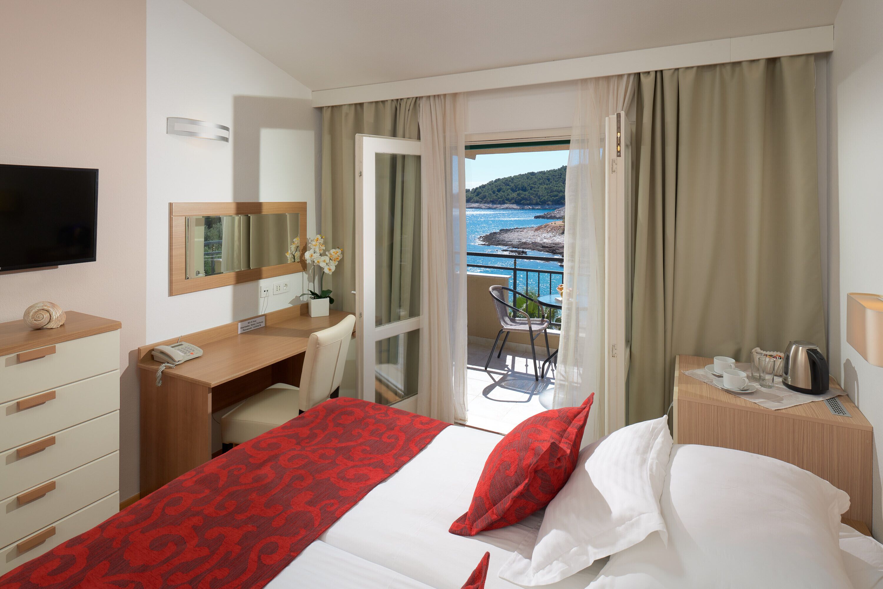 Une chambre d’hôtel comprenant un lit, un bureau, une chaise, une télévision, un balcon avec vue sur la mer et une fenêtre avec des rideaux.