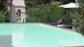 A swimming pool with lounge chairs and umbrellas, surrounded by greenery and a small tower structure.