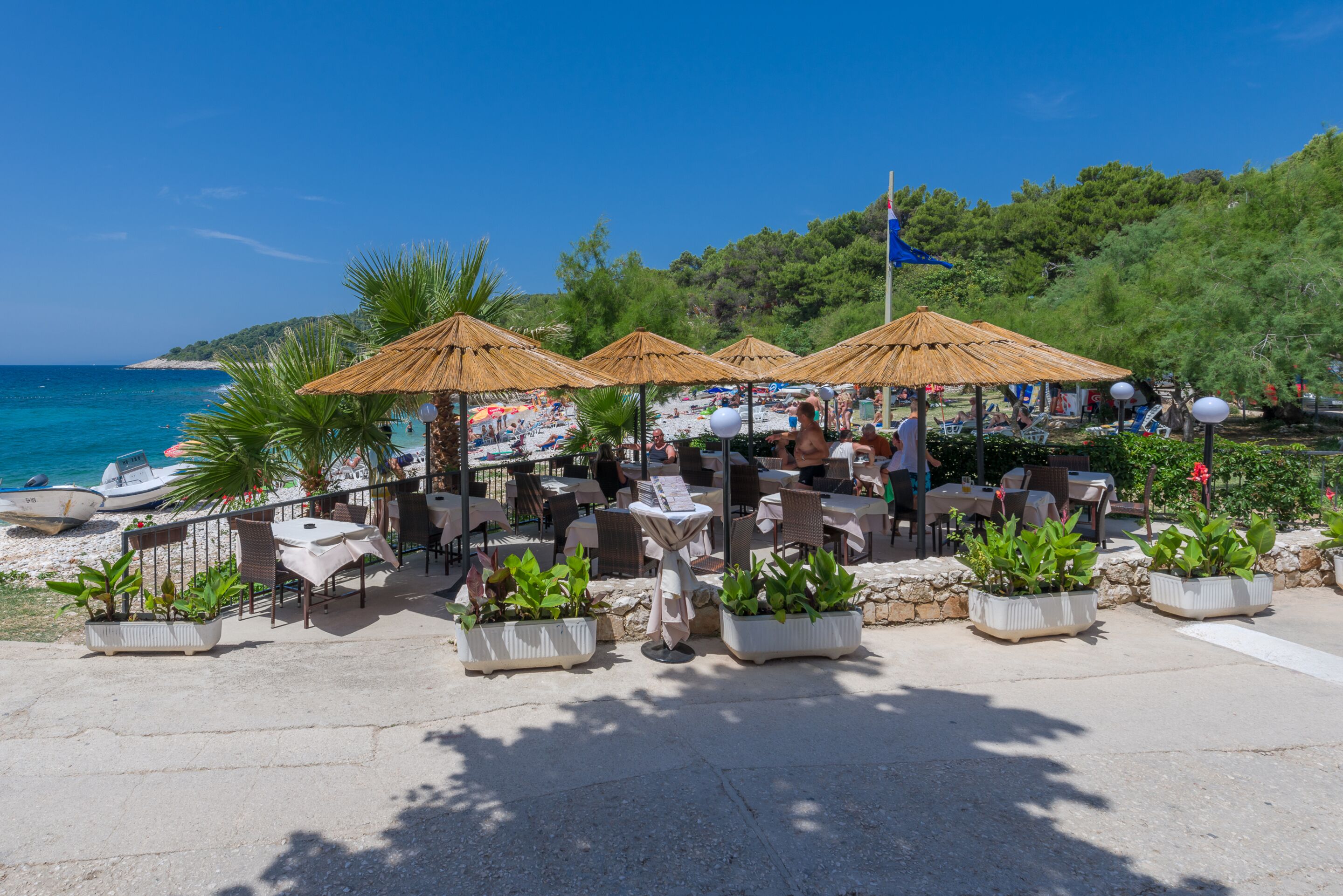 Ein Straßencafé mit Tischen und Stühlen, umgeben von Topfpflanzen und Sonnenschirmen, mit Blick auf einen Strand und das Meer.