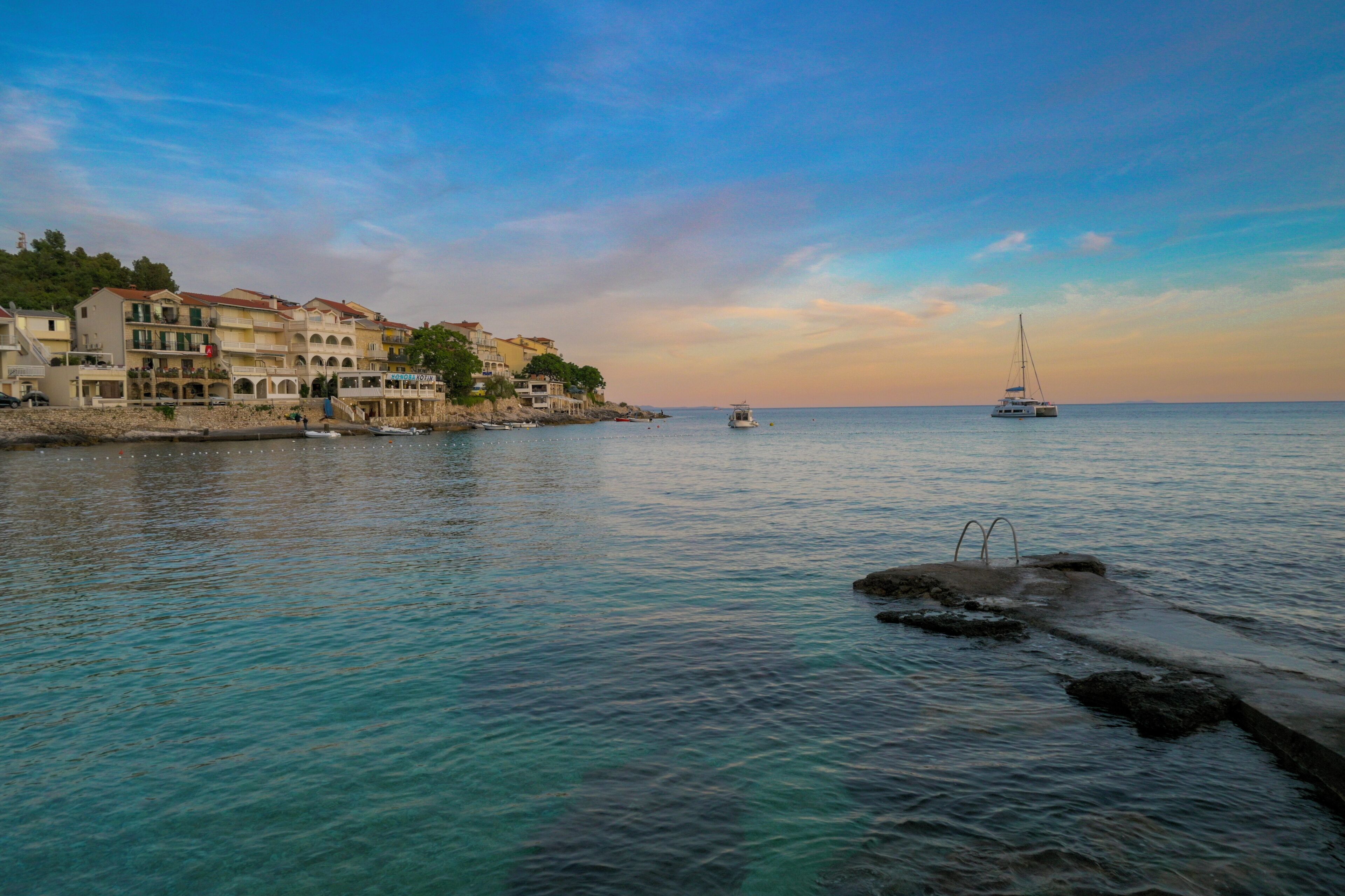 Un pueblo costero con edificios a lo largo del paseo marítimo, mar tranquilo y un velero a lo lejos.