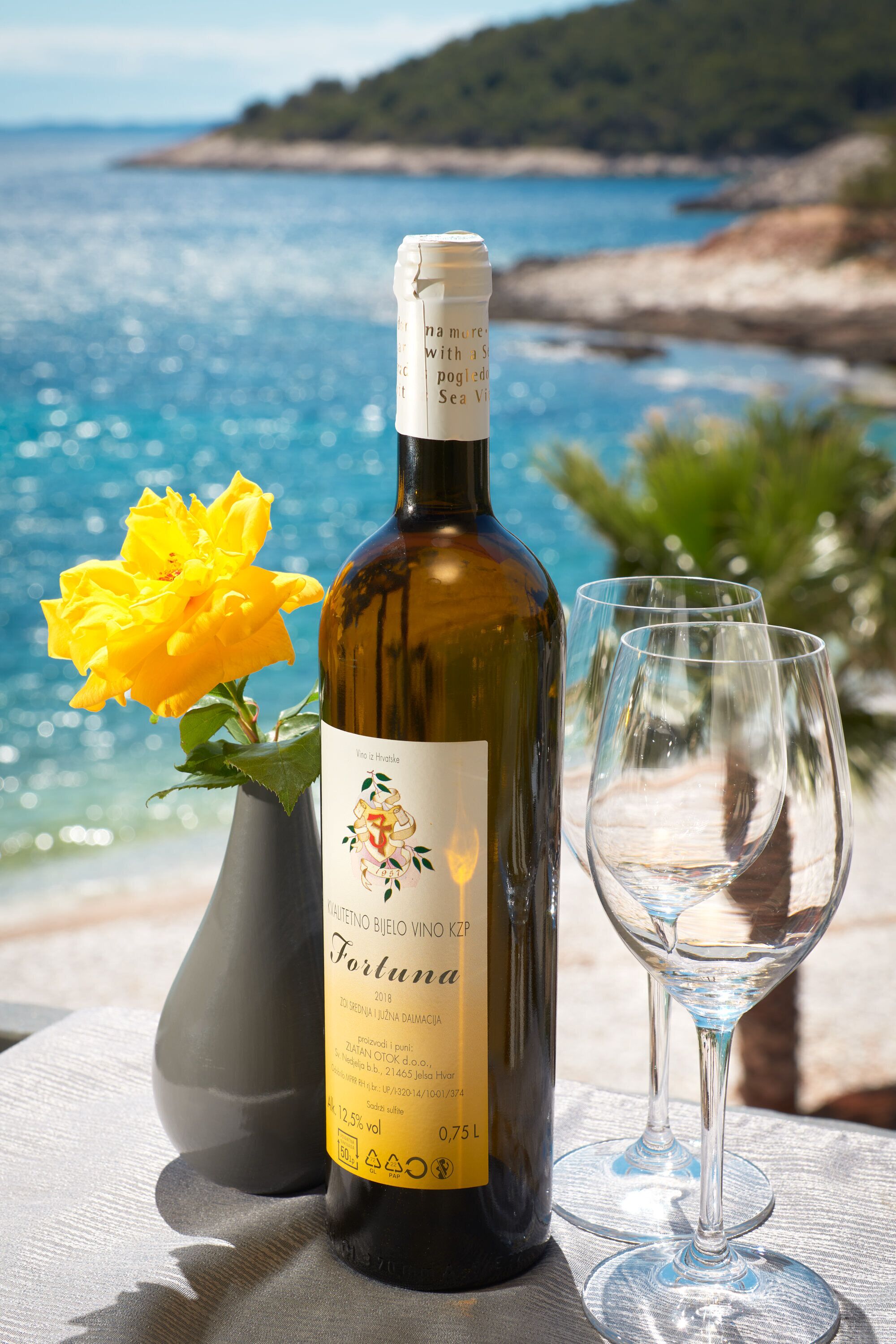 A bottle of wine with a label, a glass, and a yellow flower on a table by a scenic coastal view.