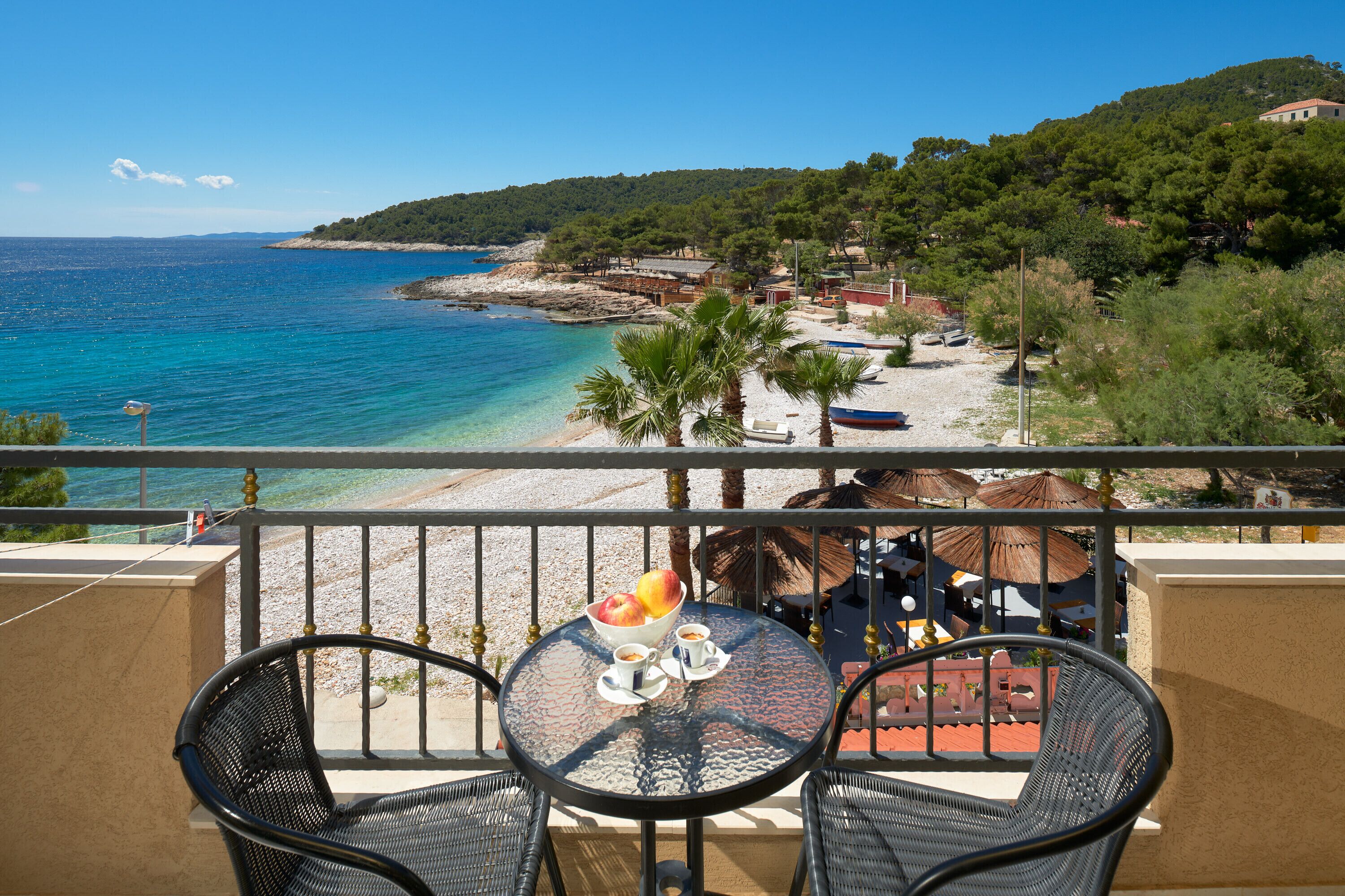 A balcony with two chairs and a table, overlooking a beach with clear blue water and a hilly landscape in the background.