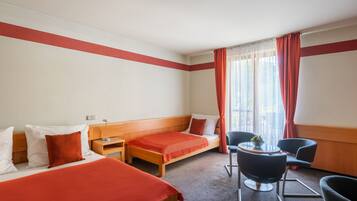 A hotel room with two single beds, a small table with a chair, and a window with curtains.