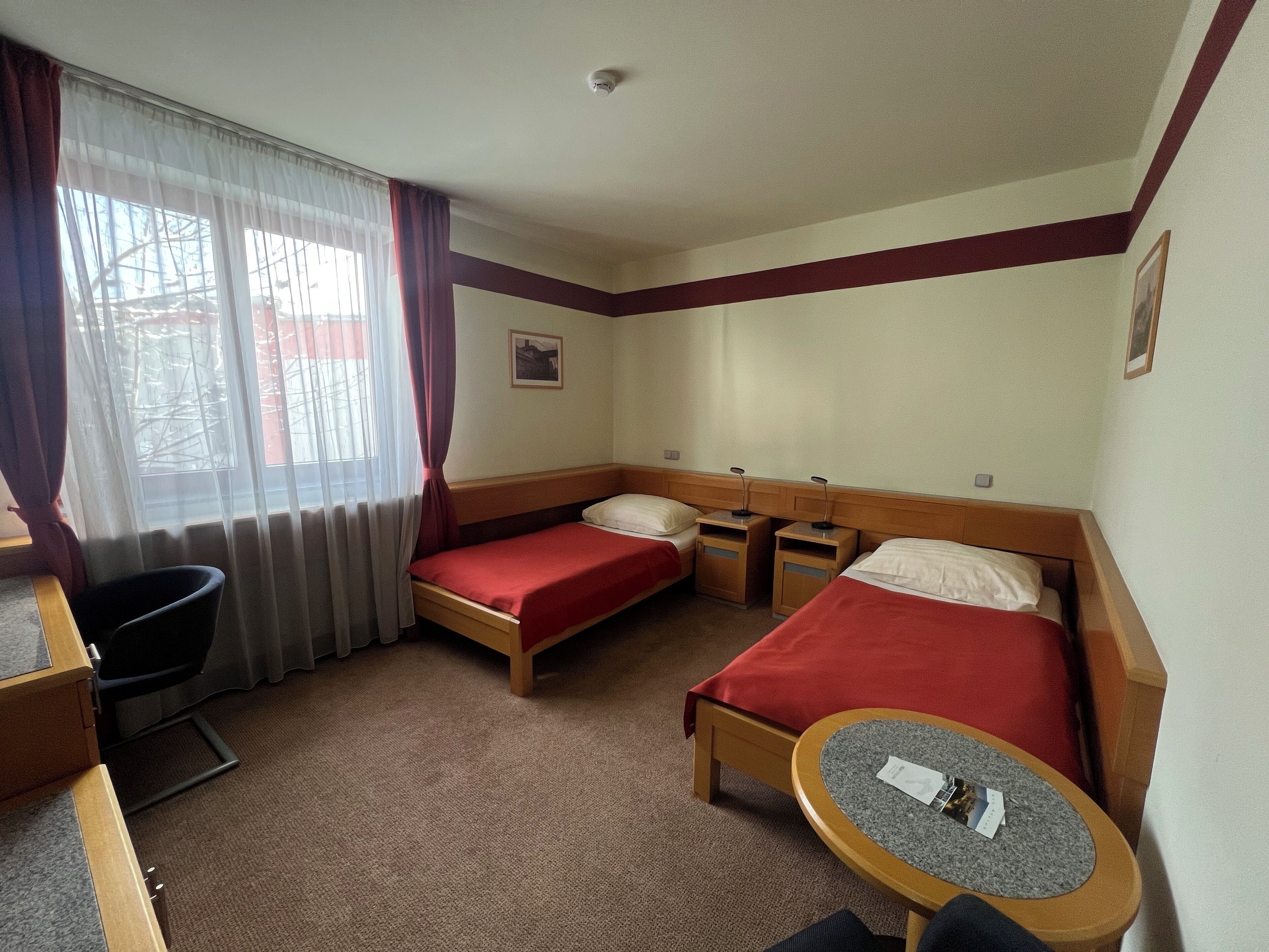A hotel room with two single beds, a desk, a chair, a small table, and a window with curtains.