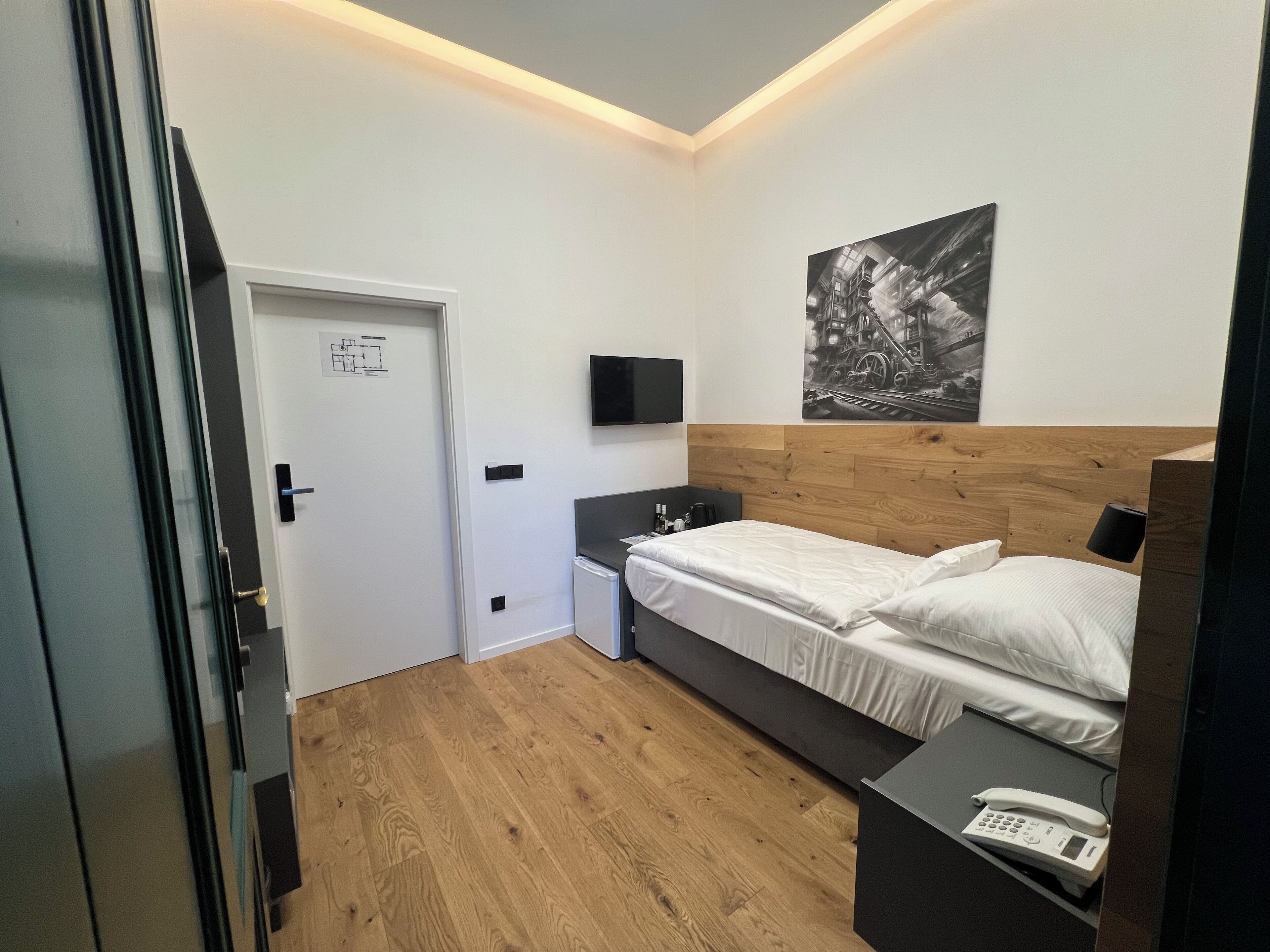 Una habitación de hotel moderna con cabecera de madera, cama con sábanas blancas, mesita de noche con teléfono y televisor montado en la pared.