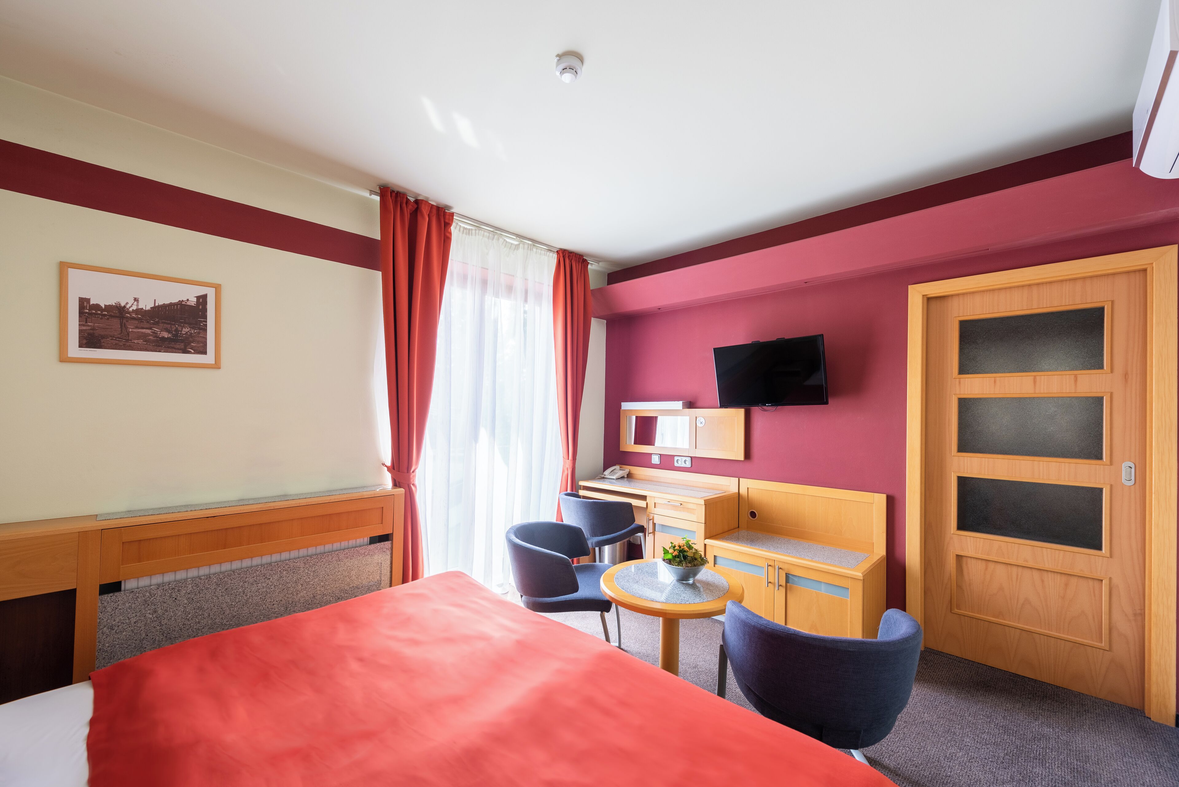 A hotel room with a bed, desk, chairs, a small table with a plant, a TV mounted on the wall, and a wooden door with frosted glass panels.