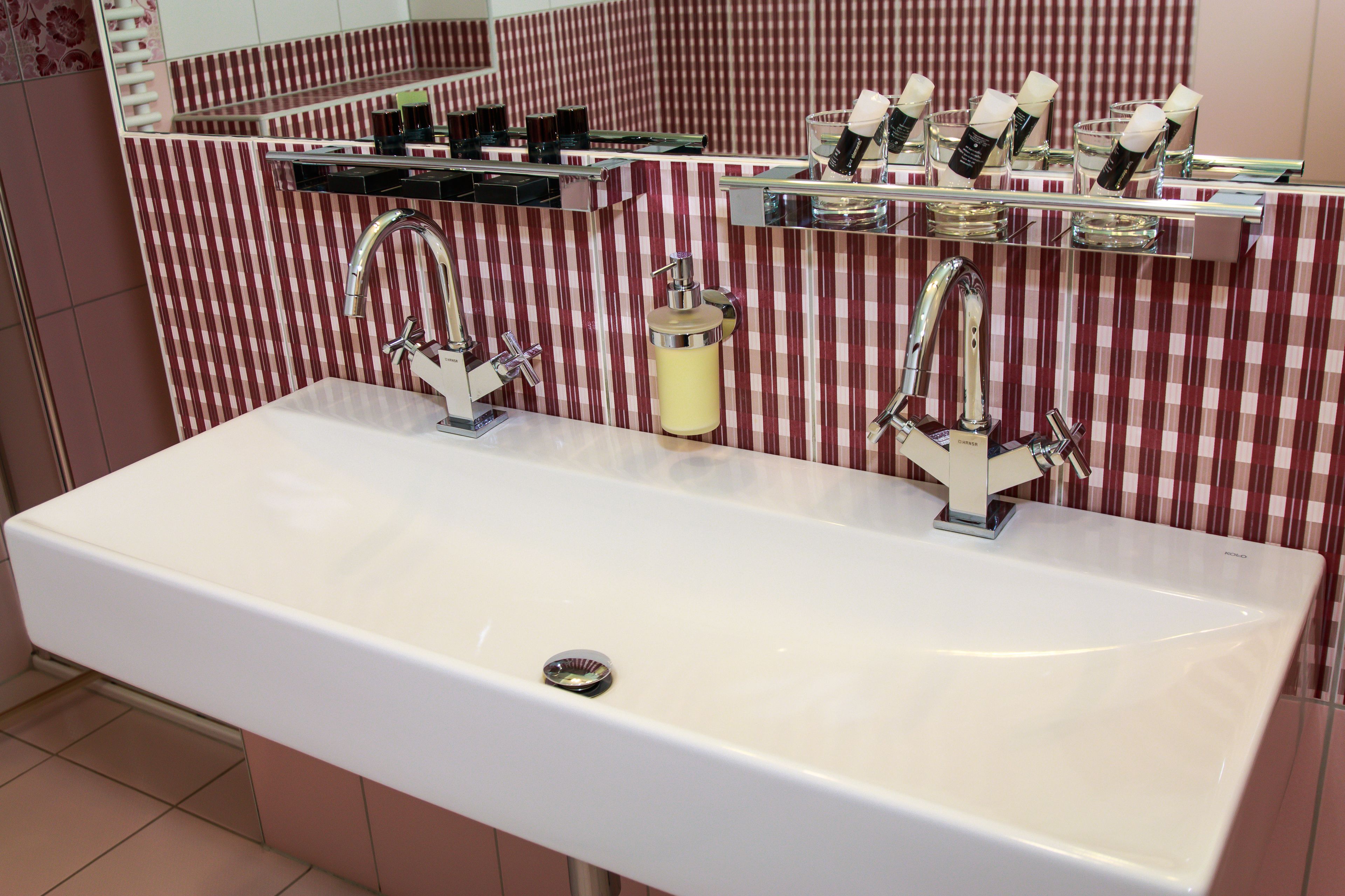 Un baño con un lavabo blanco, dos llaves y un espejo sobre el lavabo. Hay artículos de aseo en un estante sobre el espejo.